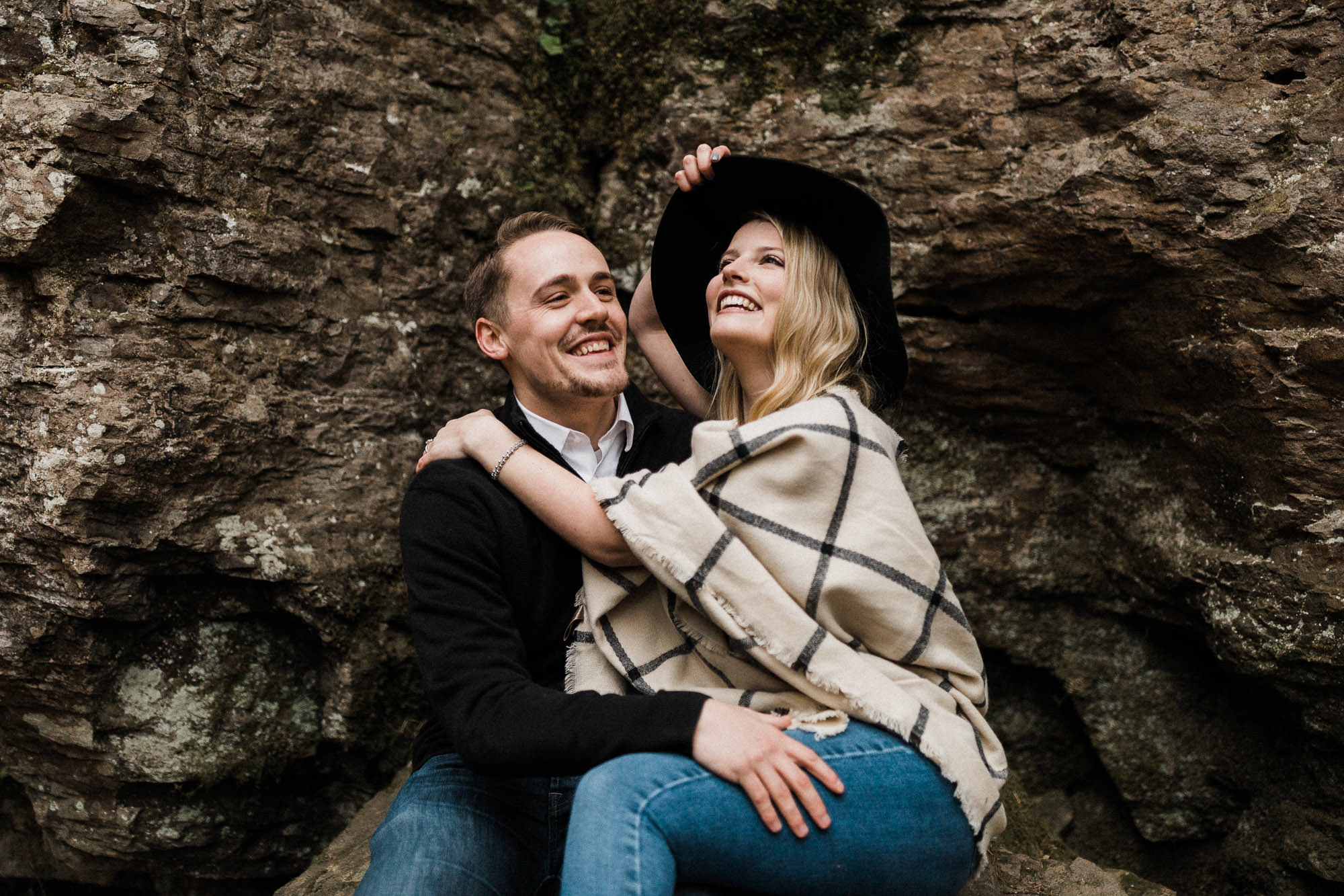 Woman sits on her fiance's lap by a rock wall in the Columbia River Gorge