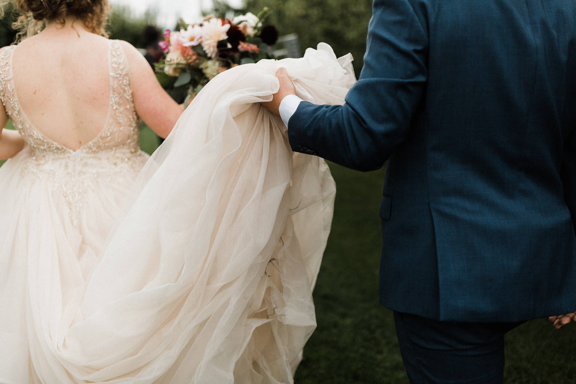 Groom holds the train of the bride's dress as they walk at Mt. View Orchards.