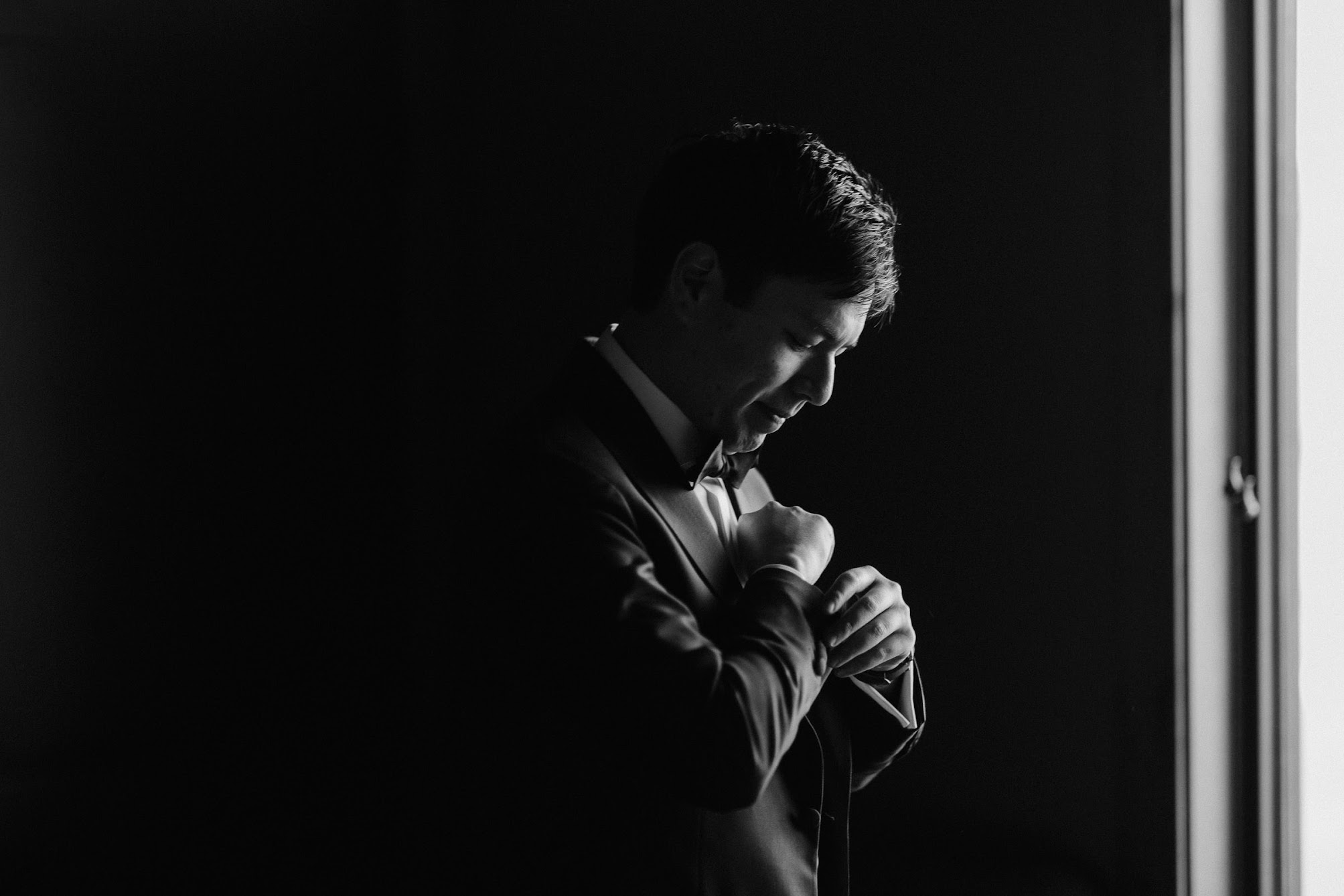 The groom looking down at his cufflinks making sure they are secured. He is wearing a suit with a collard shirt and bowtie. 