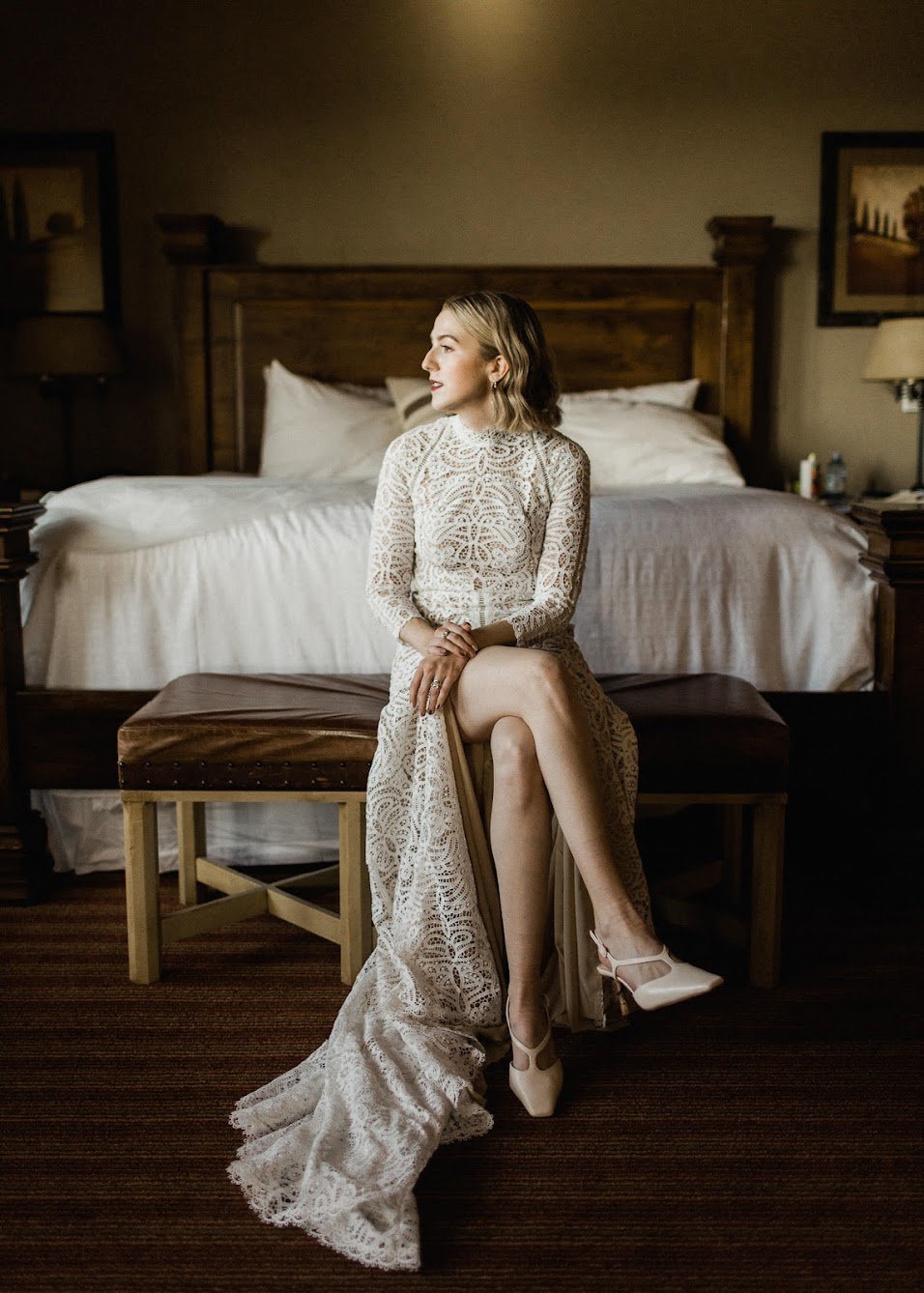 The bride sitting on a leather bench, looking out the window with her legs crossed. Her wedding dress has a classy slit so you can see her legs, and her stunning close-toes heels!