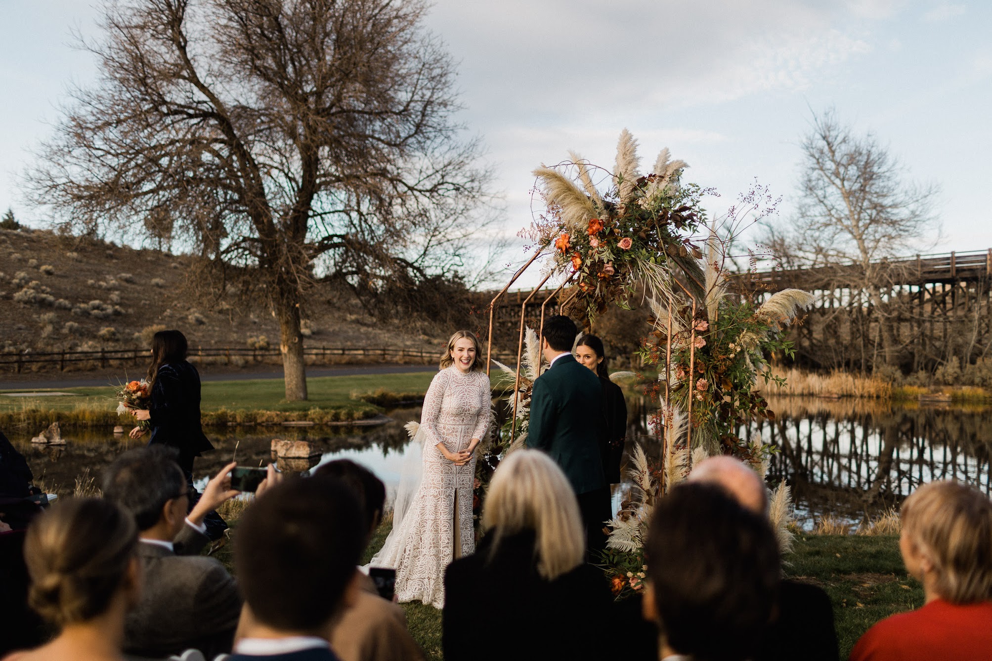 Bride and groom standing at the arbor with their officiant. The bridesmaid of honor is walking away with her bridal bouquet. The ceremony has started.