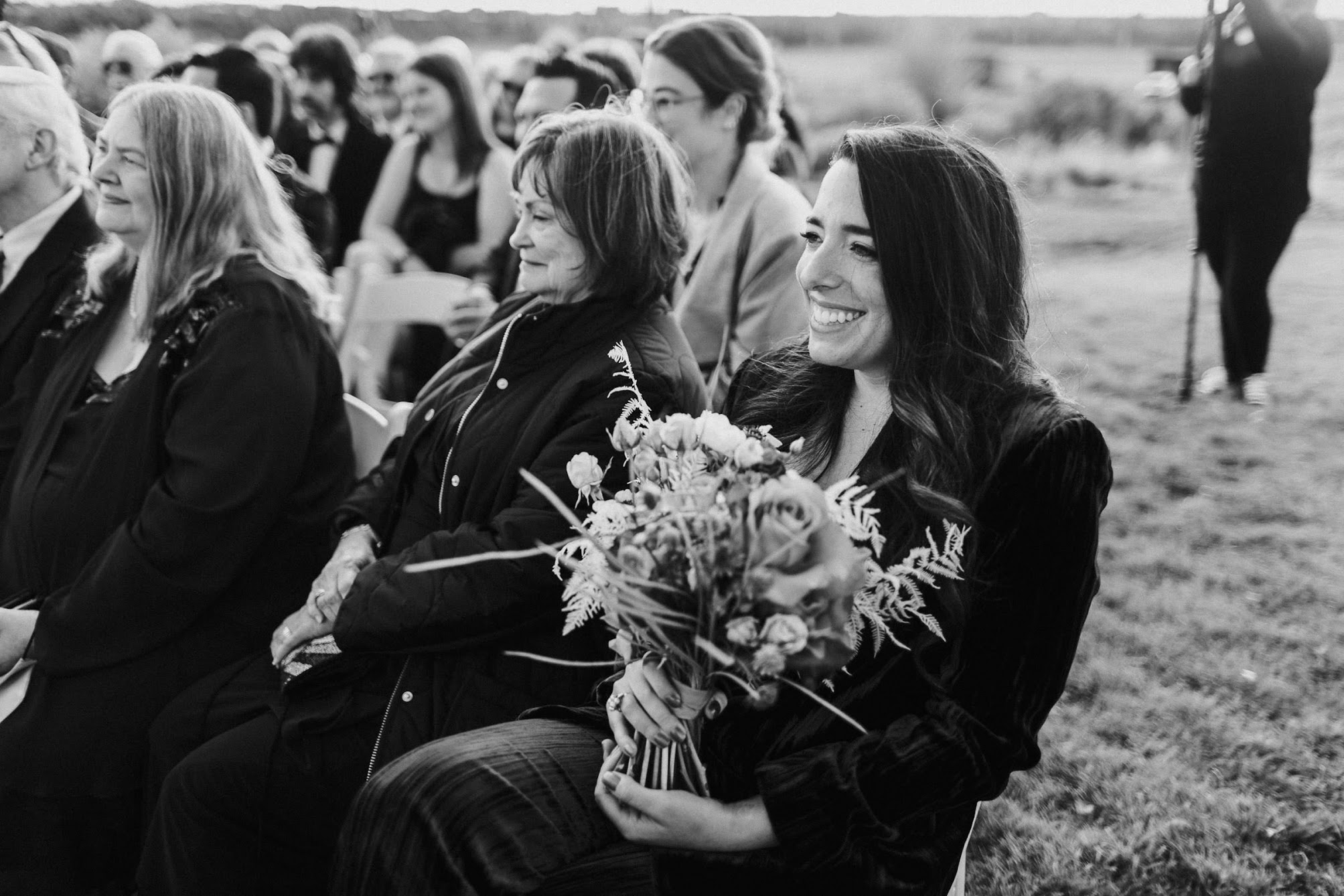 Maid of honor is smiling holding the bridal bouquet as she watches the ceremony. 