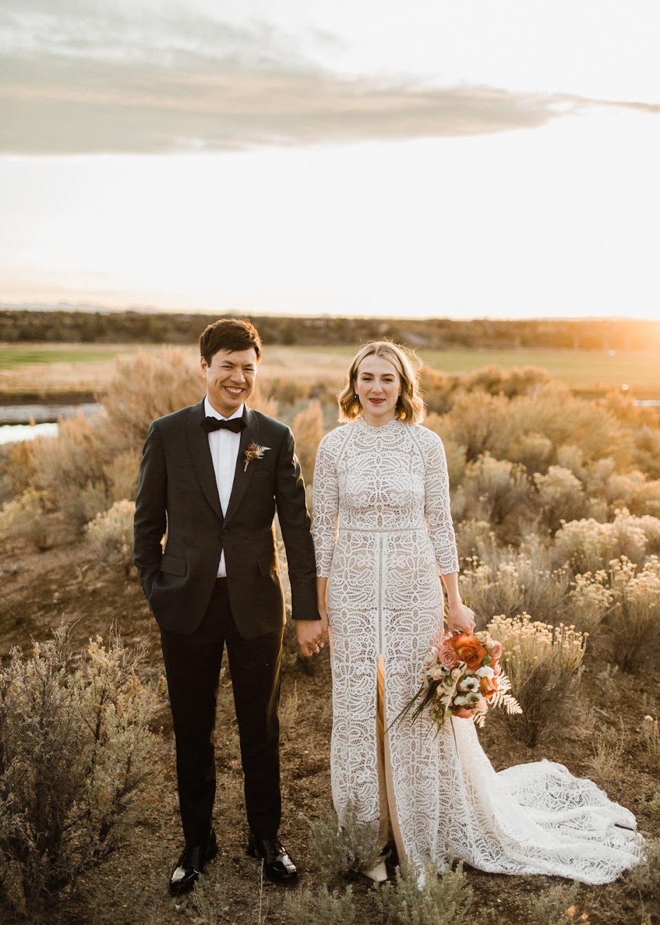 the bride and groom standing side by side hold hands looking into the camera 