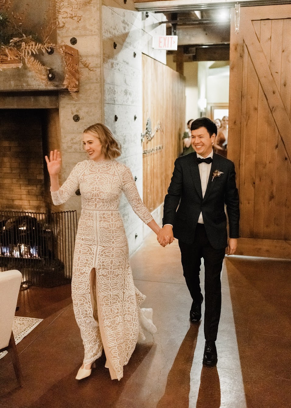 the bride and groom walking into the wedding reception hold hands, smiling and waving at their guests. 