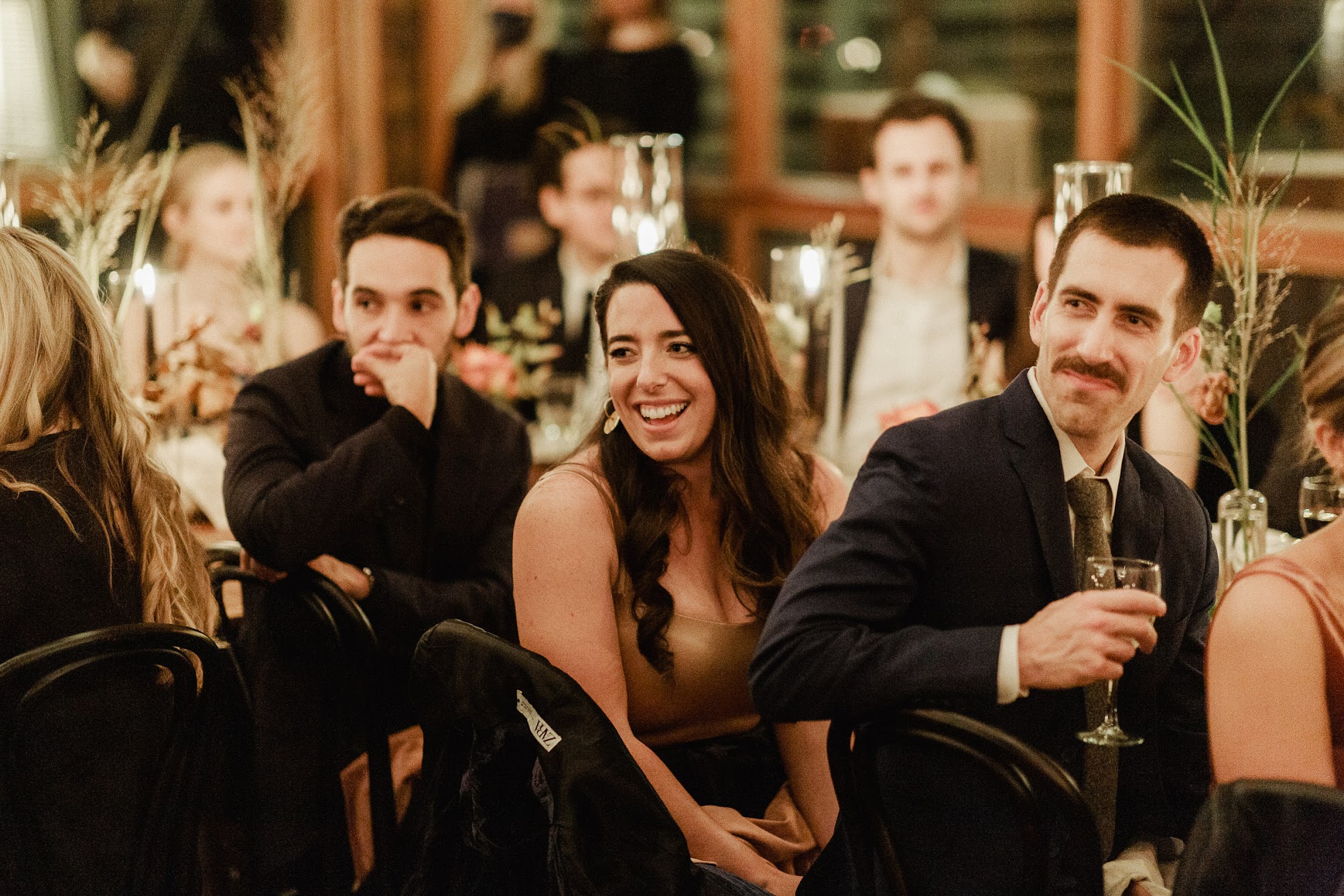 Guests have a drink and their hands and are laughing while the mother of the bride tells a funny story