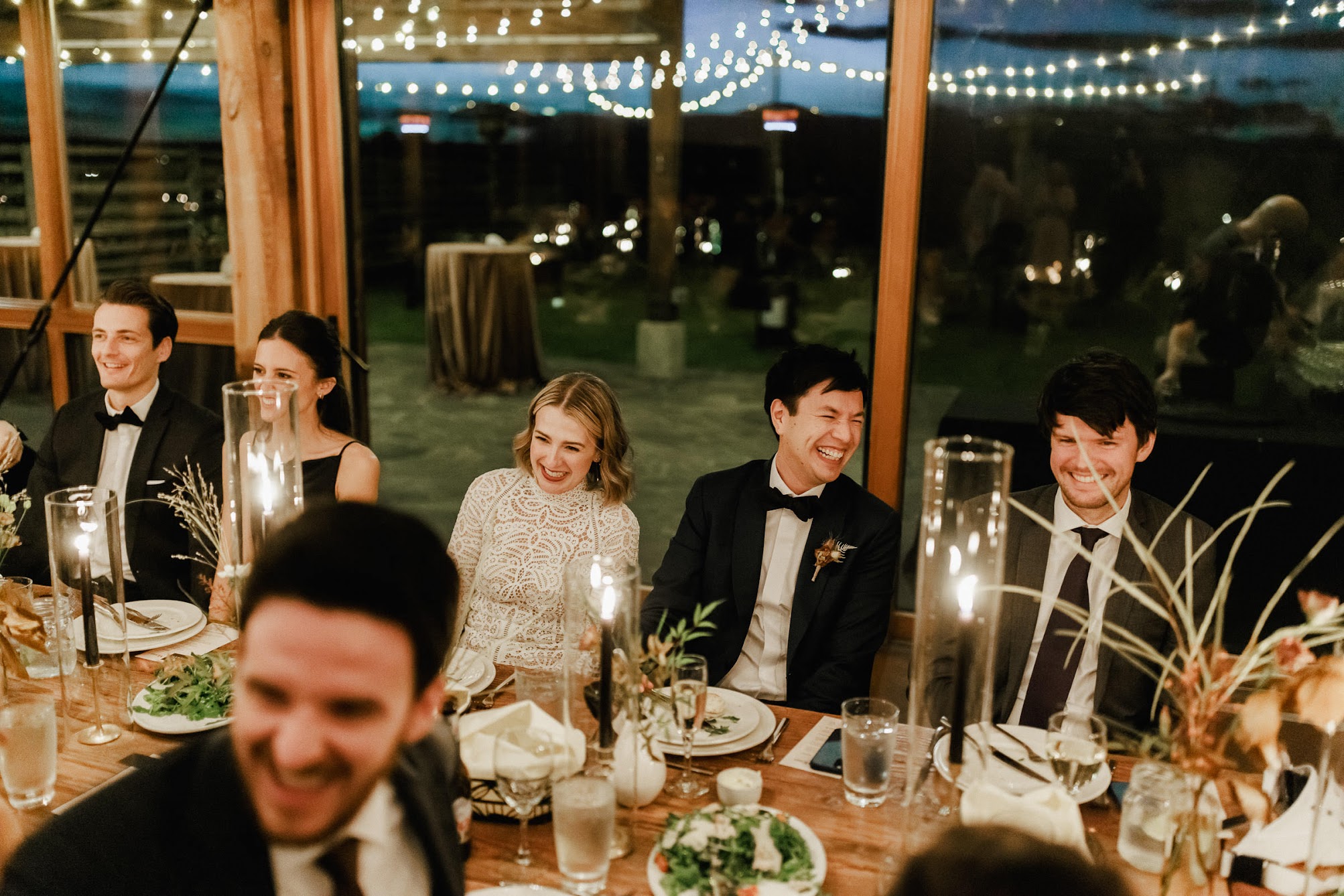 The bride and groom are sitting at their table laughing. Behind them you see string of bistro lights.