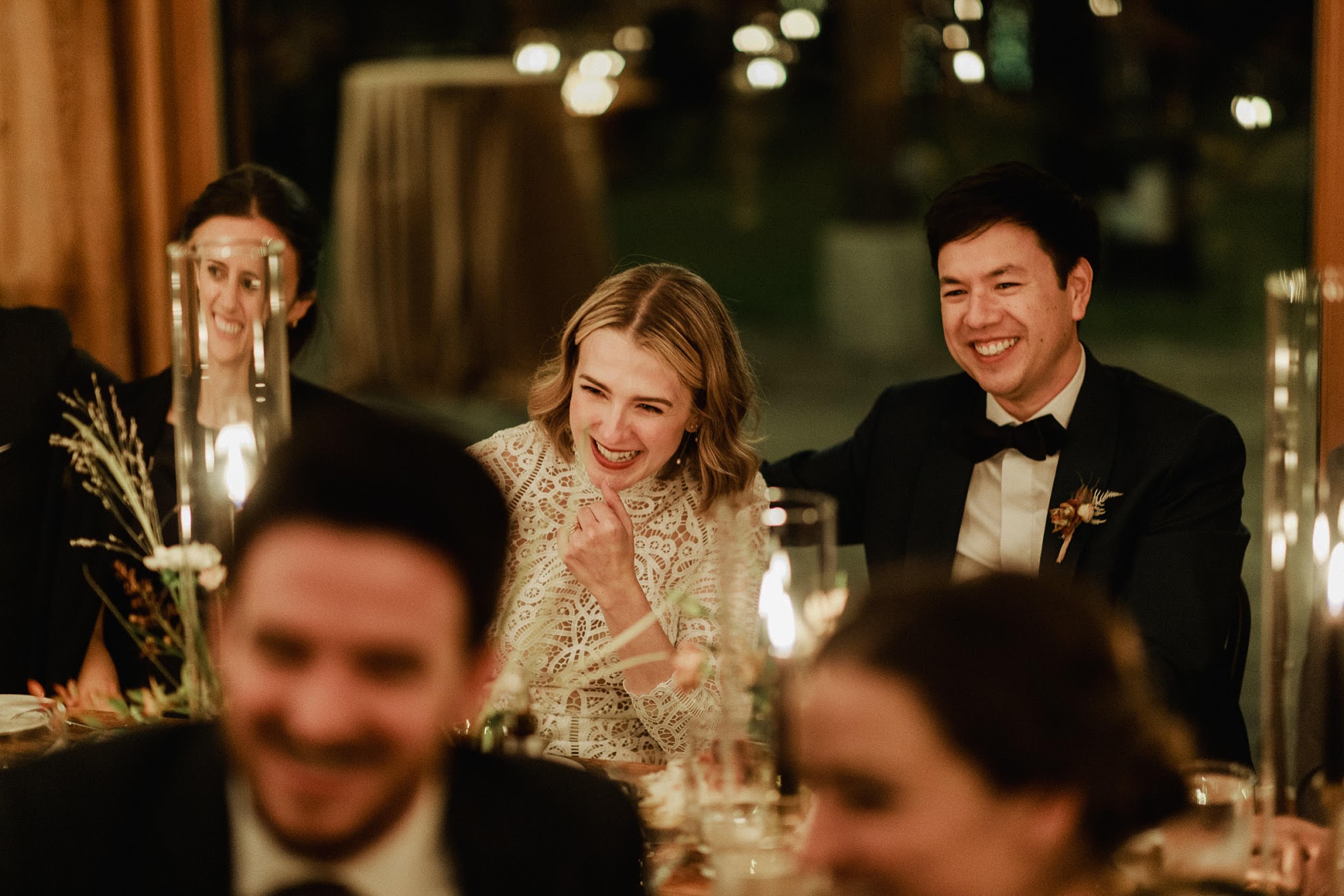 The bride and groom sitting at their dinner table, surrounded by guests, all laughing.