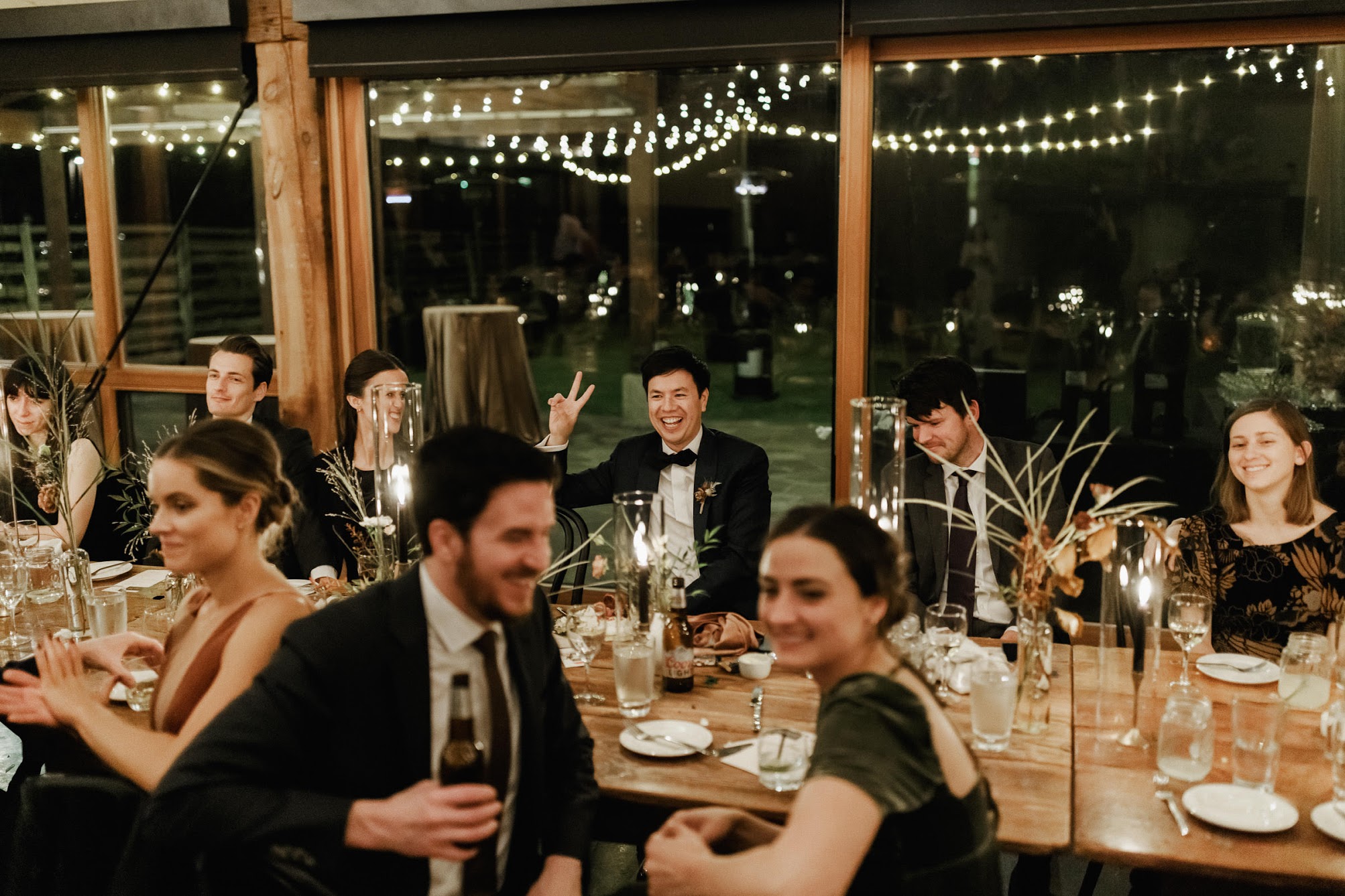 The groom sitting at the table holds up the peace sign, laughing while his wife is giving a speech.