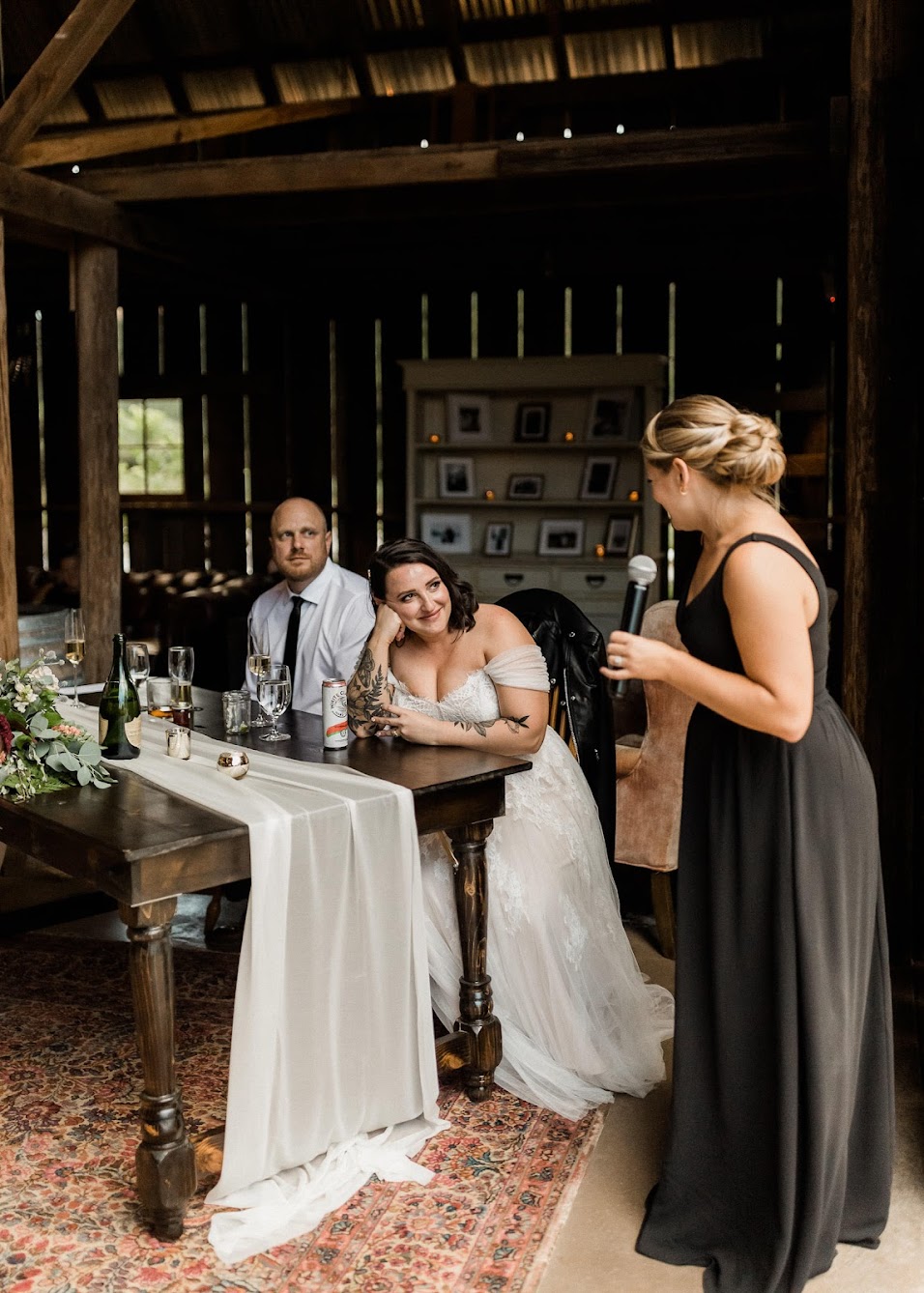 The maid of honor standing next to the sweetheart table giving a speech, the bride is looking at her and smiling. 