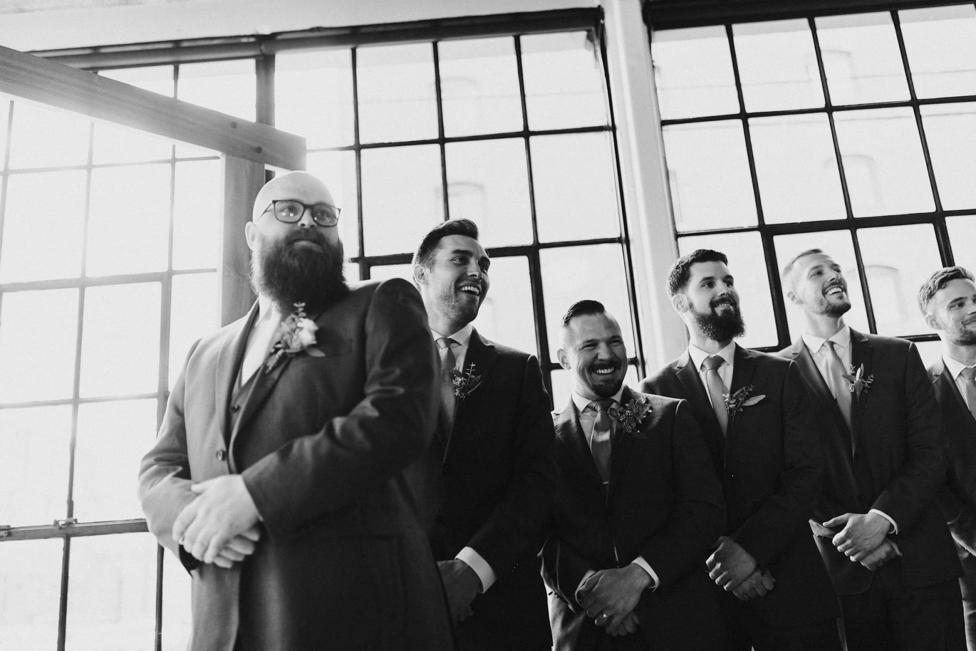 the groomsmen and groom watching the bride walk towards the arbor 