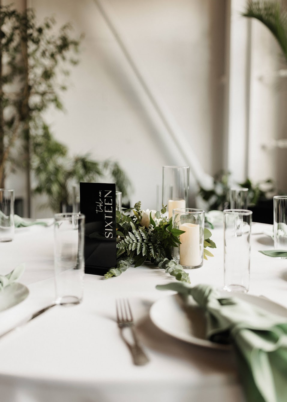 a wedding reception table with greenery as a centerpiece, a modern table number sign and pillar candles 