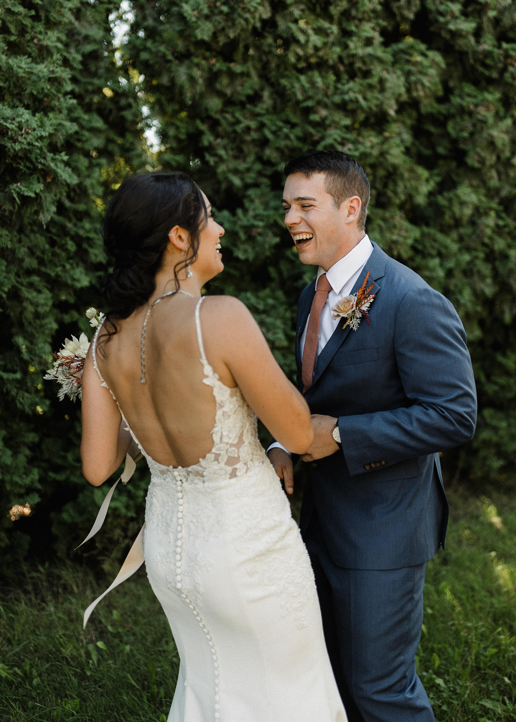 Bride and groom laugh during a first look in front of a row of hedge trees