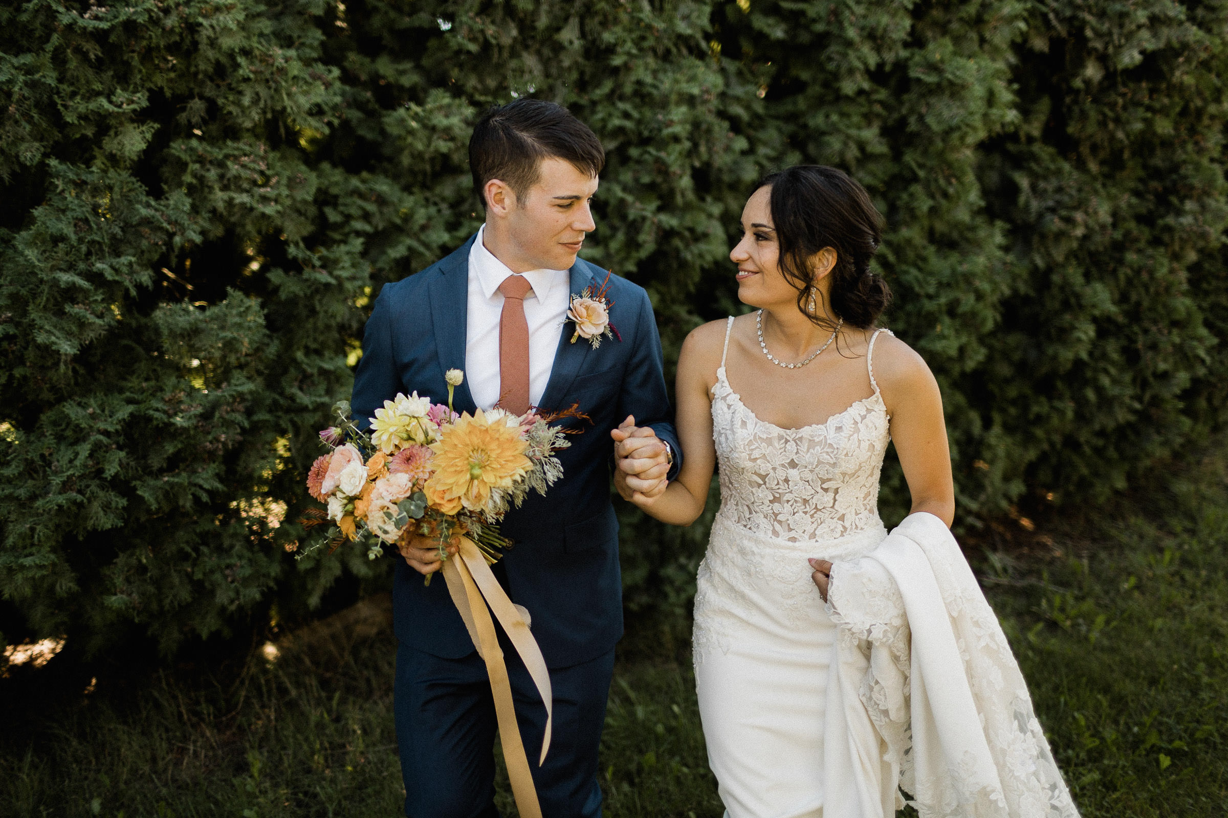 Bride and groom hold hands during a first look in front of a row of hedge trees