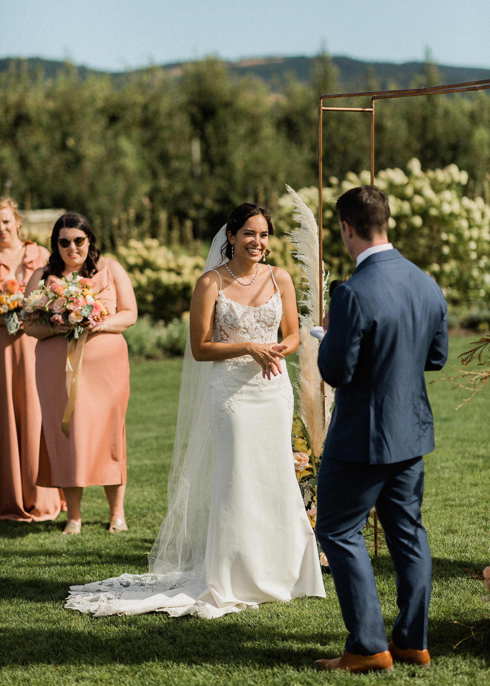 Bride smiles as groom reads vows during wedding ceremony at The Orchard