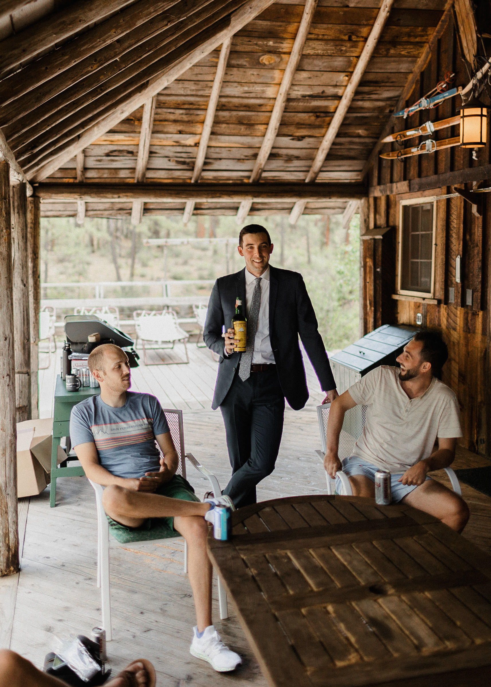Groom, standing on a deck outside a cabin, holds a bottle of scotch as his groomsmen look on