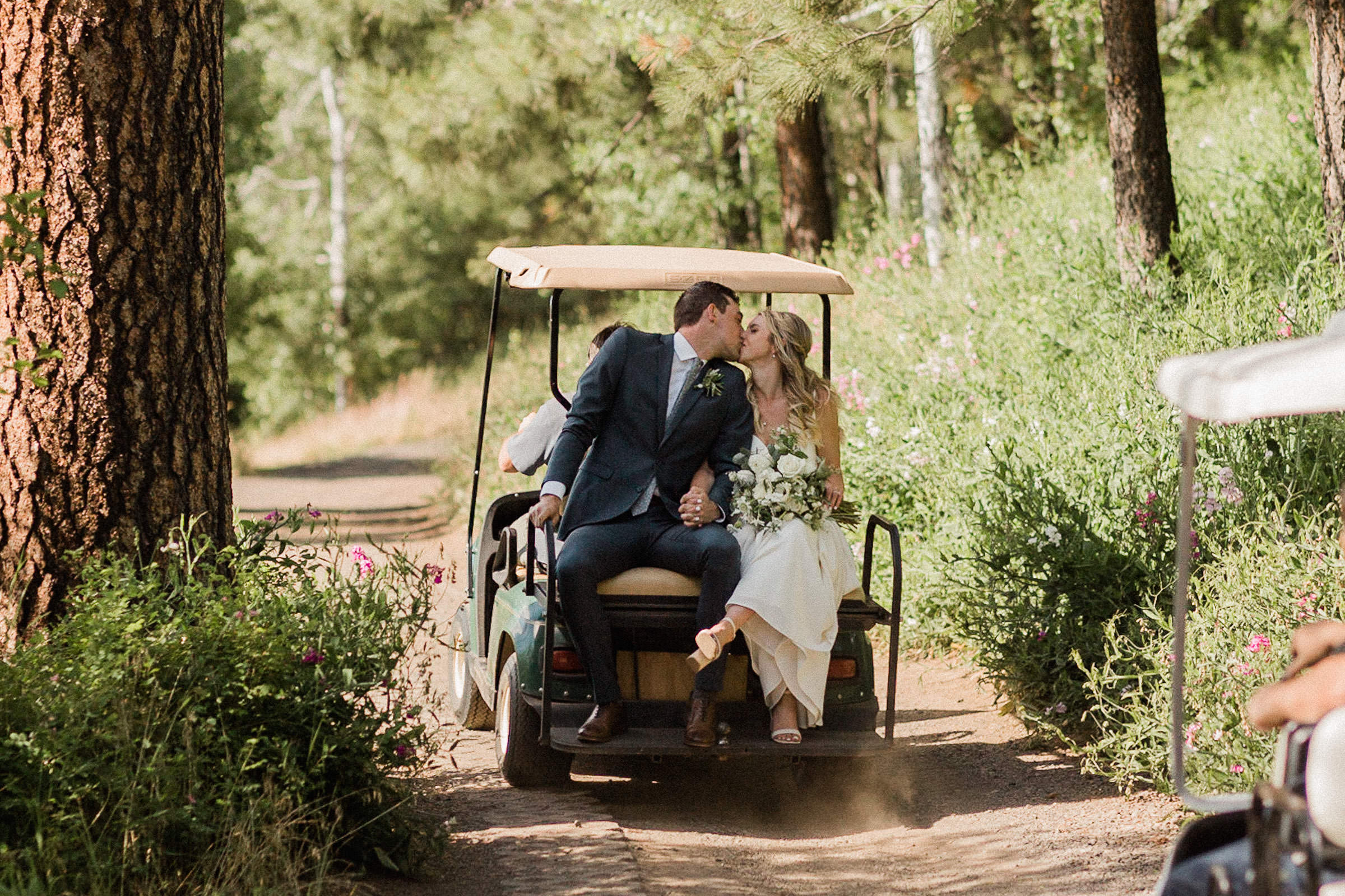 Bride and groom kiss while being driven away on the back of a golf cart